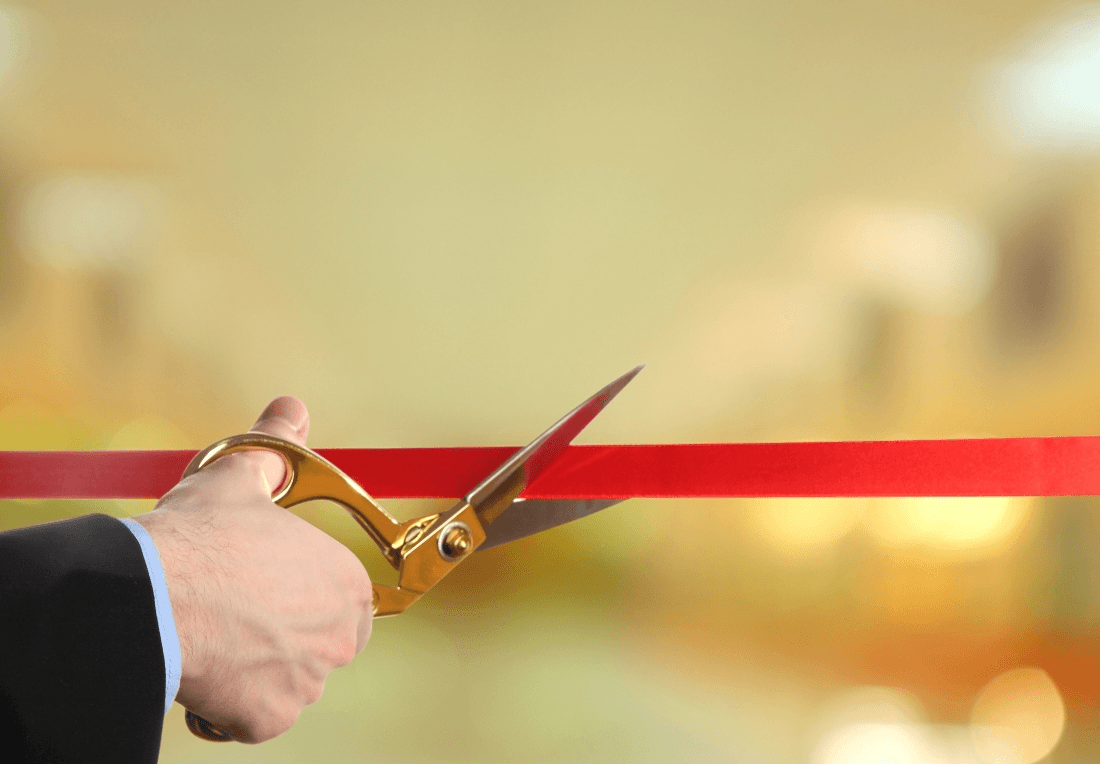 A hand in a dark suit uses gold-handled scissors to cut a red ribbon, marking a grand opening against a softly glowing gold background.