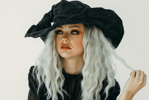 A woman in a dramatic black witch hat and dark Halloween clothing gazes upward, her silver-white hair framing smoky eye makeup and a contemplative expression.