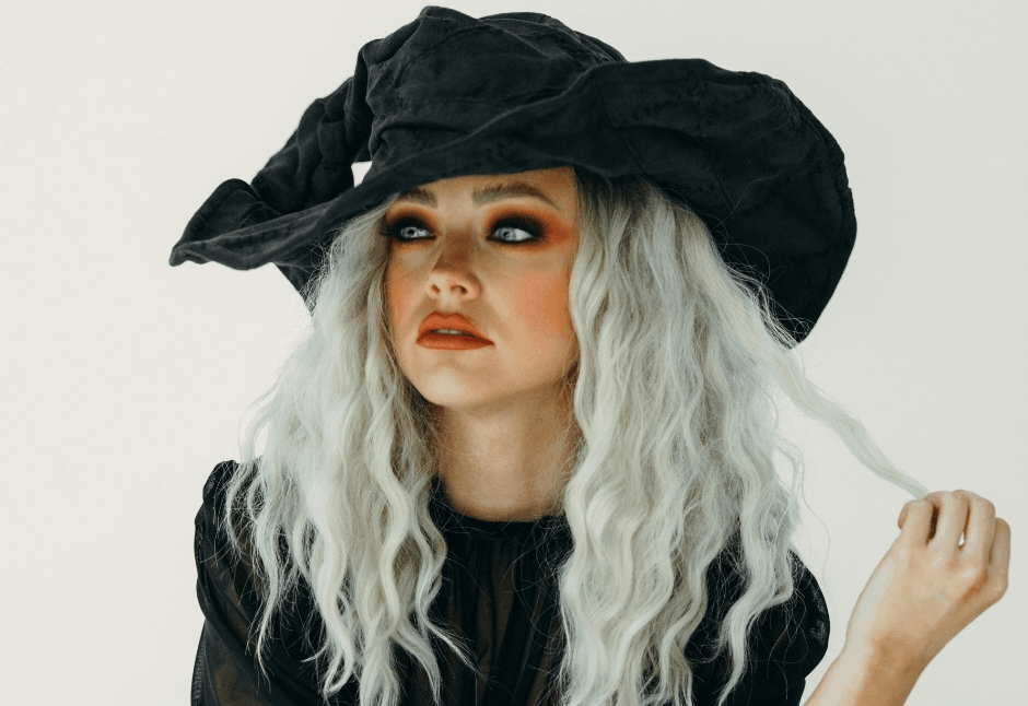 A woman in a dramatic black witch hat and dark Halloween clothing gazes upward, her silver-white hair framing smoky eye makeup and a contemplative expression.
