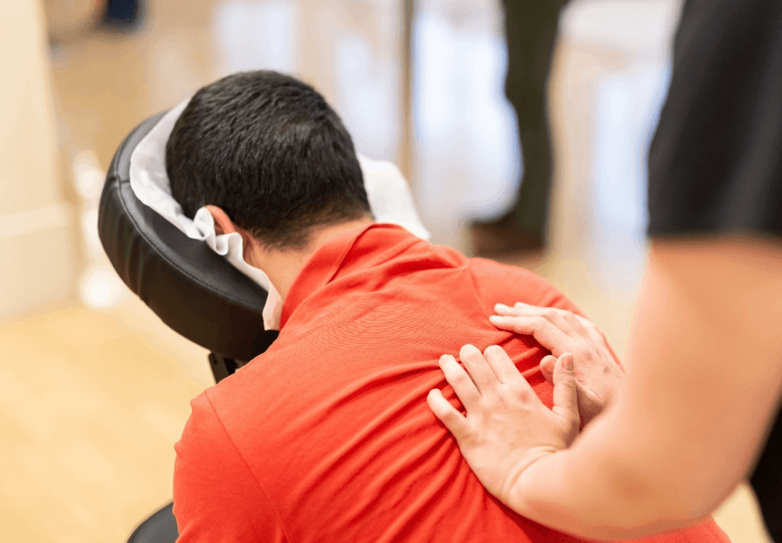 A person in a red shirt leans forward in a chair massage as a therapist gently presses their shoulders, creating a calm, relaxing wellness scene.