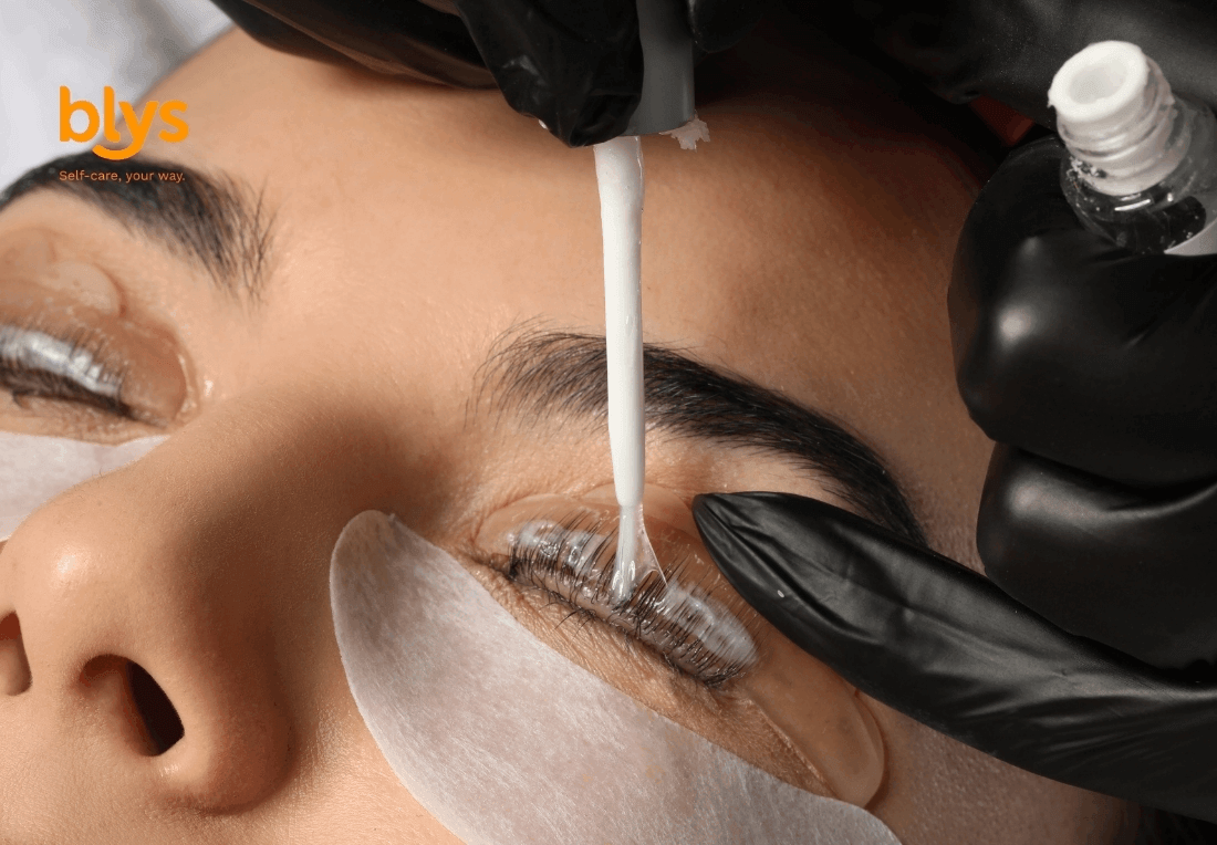 Close-up of a woman receiving an eyelash tint treatment as a gloved technician applies product to her lashes above an under-eye patch; calm beauty-care scene.