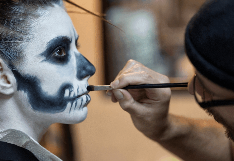 A makeup artist paints skeletal teeth on a person’s cheek, completing dramatic blue-and-white skeleton makeup in a focused backstage scene.