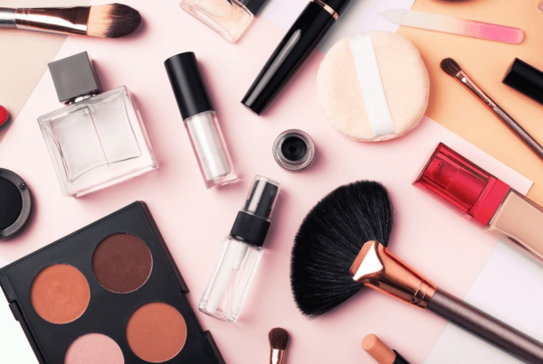 Flat lay of makeup brushes, palettes, lipstick, mascara, perfume, and other cosmetics on a pale pink background, creating a polished, playful beauty mood.