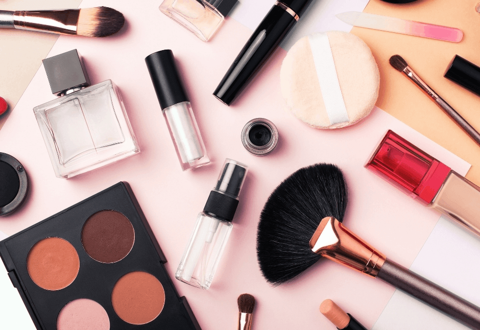 Flat lay of makeup brushes, palettes, lipstick, mascara, perfume, and other cosmetics on a pale pink background, creating a polished, playful beauty mood.