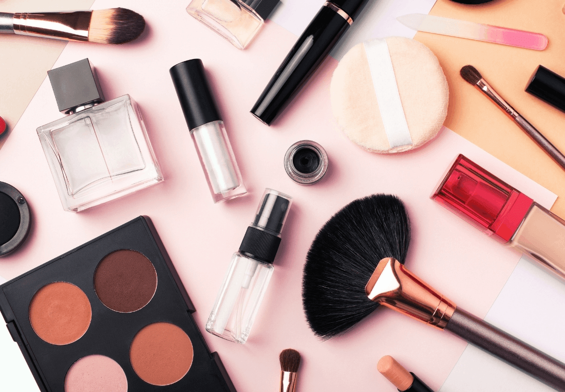 Flat lay of makeup brushes, palettes, lipstick, mascara, perfume, and other cosmetics on a pale pink background, creating a polished, playful beauty mood.