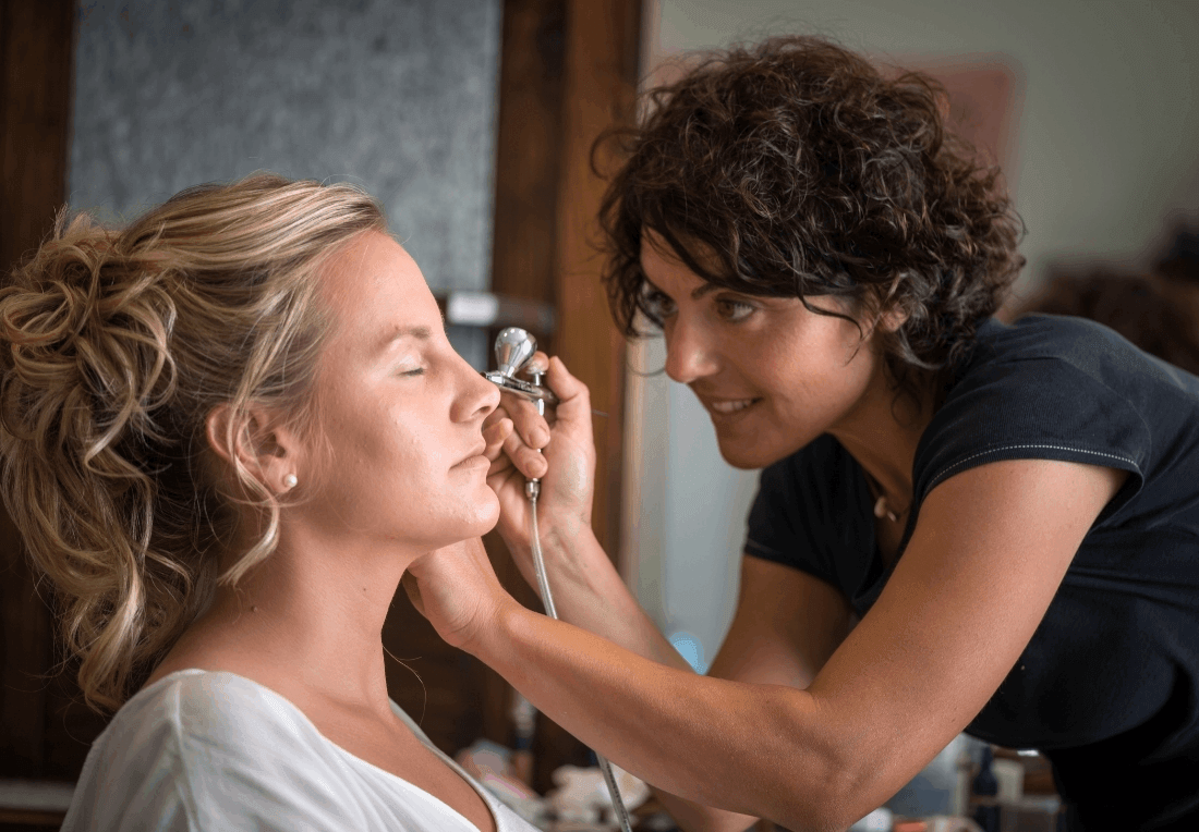 A makeup artist gently applies airbrush makeup to a seated blonde woman, who keeps her eyes closed; their close interaction feels calm and focused.