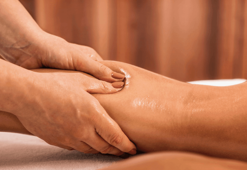 A therapist gently massages an oil-sheened client’s lower lipoedema legs on a treatment table, with warm curtains behind, creating a calm atmosphere.