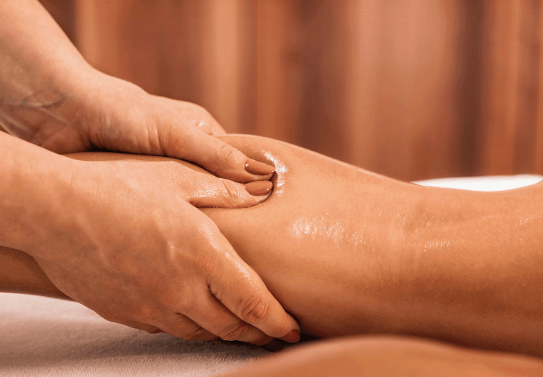 A therapist gently massages an oil-sheened client’s lower lipoedema legs on a treatment table, with warm curtains behind, creating a calm atmosphere.