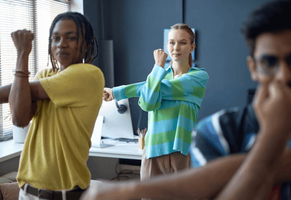 Three coworkers in an office stretch their arms beside a sunlit window, with a woman in a green-and-blue striped sweater centered, projecting a relaxed, upbeat corporate wellness lunch and learn mood.