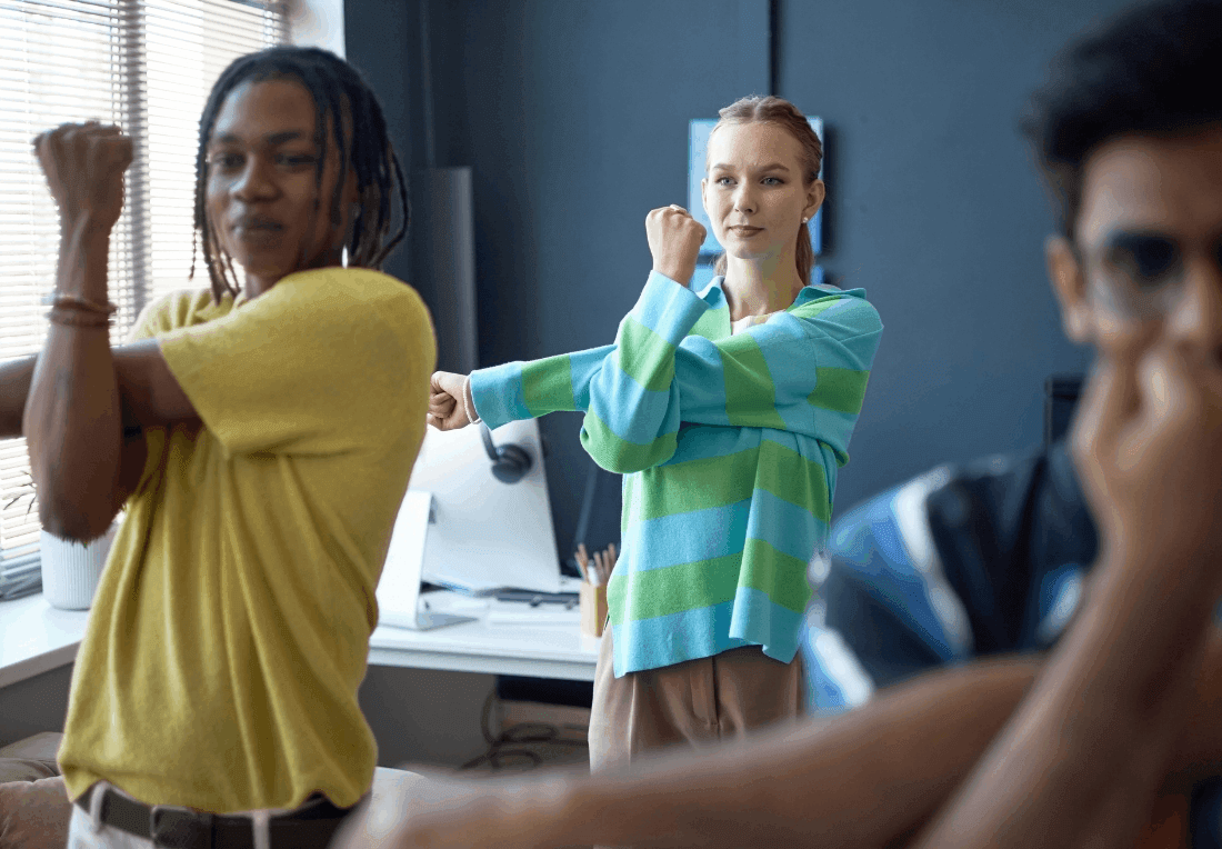 Three coworkers in an office stretch their arms beside a sunlit window, with a woman in a green-and-blue striped sweater centered, projecting a relaxed, upbeat corporate wellness lunch and learn mood.