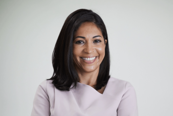 A smiling woman with shoulder-length dark hair wears a pale lavender blouse in a corporate headshot against a soft gray background.