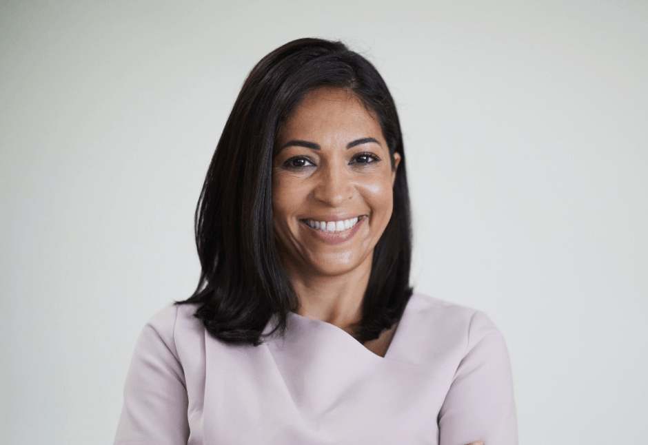 A smiling woman with shoulder-length dark hair wears a pale lavender blouse in a corporate headshot against a soft gray background.