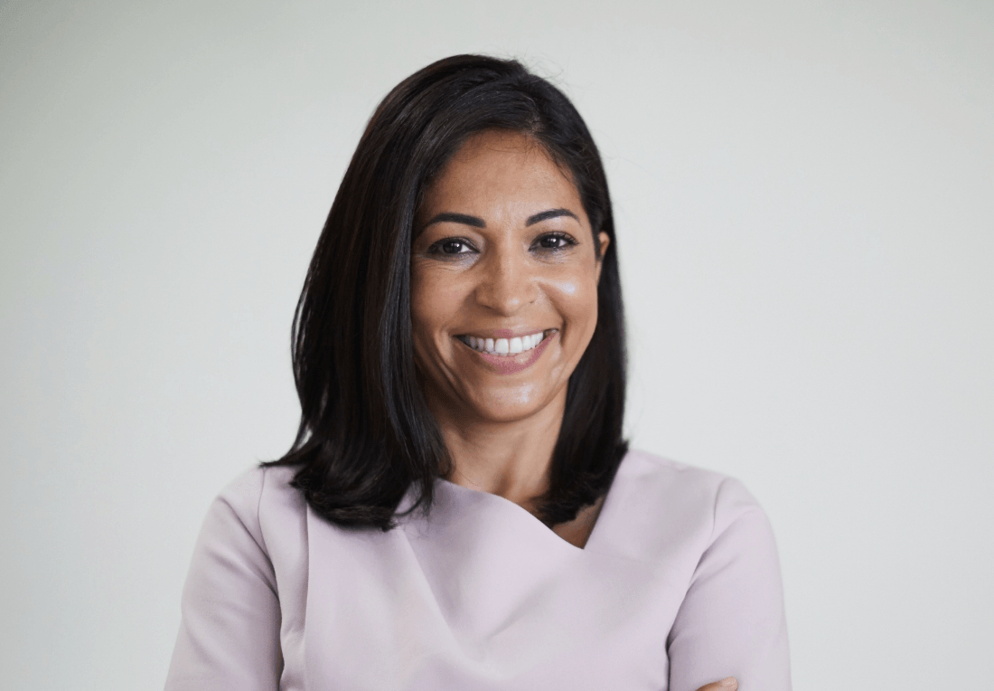 A smiling woman with shoulder-length dark hair wears a pale lavender blouse in a corporate headshot against a soft gray background.