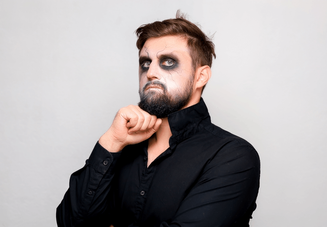 Bearded man in a black shirt poses against a plain gray background with dark, smudged eye makeup, chin resting on his fist and a gothic Halloween look.