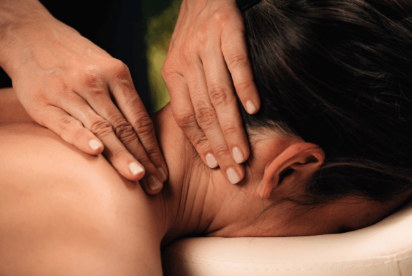 Close-up of hands gently massaging a woman’s neck and shoulder as she lies face down on a massage table, conveying calm and relaxation from forward head posture, rounded shoulders and neck hump from sitting at work.