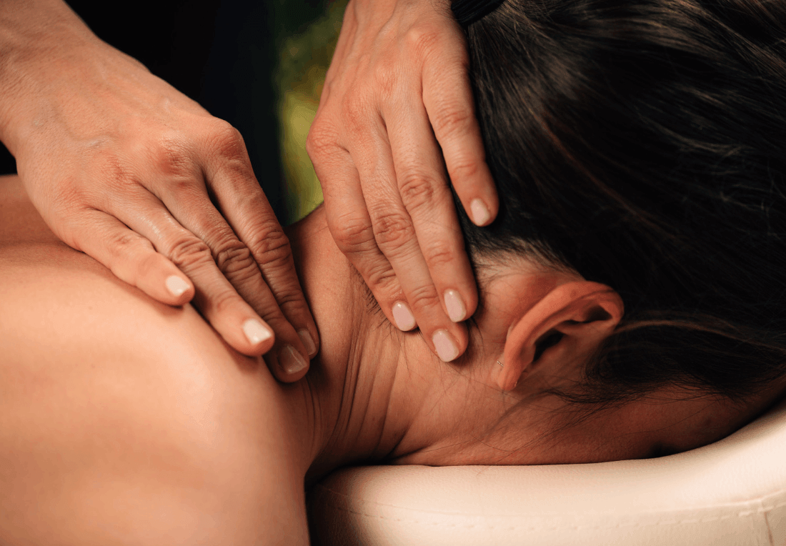 Close-up of hands gently massaging a woman’s neck and shoulder as she lies face down on a massage table, conveying calm and relaxation from forward head posture, rounded shoulders and neck hump from sitting at work.